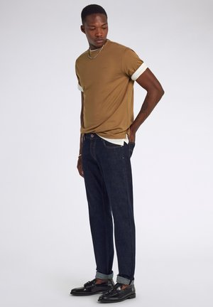 Hombre con camisa marrón de manga corta sobre una camiseta blanca, vaqueros azul oscuro remangados y mocasines negros con borlas, de pie con una mano en el bolsillo.