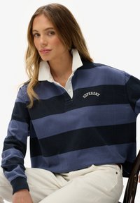 Jersey de rayas en azul marino y azul claro con cuello blanco. Presenta un logo bordado y mangas largas, mostrado en un modelo sentado.