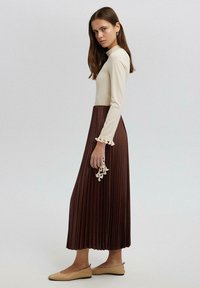 Women’s long dress featuring a fitted cream top, pleated brown skirt, and lace detailing at sleeve cuffs. Brown flats complete the outfit.