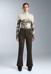 Béžový zipsový top so vzorom hôr, kombinovaný s tmavohnedými rozšírenými nohavicami s dvoma zipsovými vreckami a textúrovanými akcentmi.