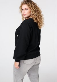Vrouw met krullend blond haar, gekleed in een zwarte trui en lichtgrijze jeans, die over haar schouder kijkt tegen een simpele witte achtergrond.