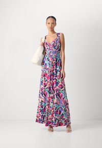 Abito lungo floreale con corpetto aderente, stampa di foglie multicolori su sfondo chiaro, realizzato in tessuto leggero, abbinato a una borsa bianca.