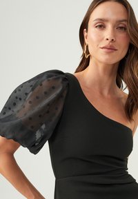 Black dress with a fitted silhouette and one shoulder design. Puff sleeve made of sheer fabric with black polka dots. Smooth texture.