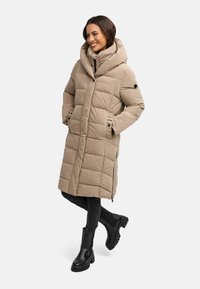 Cappotto lungo beige trapuntato con un grande cappuccio e cerniere laterali, abbinato a jeans neri e stivali neri chunky. Materiale liscio e design elegante.