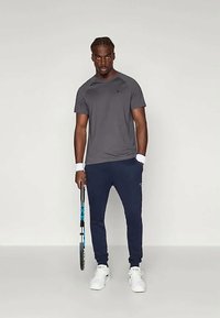 Grå treningstopp og marineblå joggebukser, kombinert med hvite sneakers. Holder en tennisracket. Enkel design med en avslappet passform.