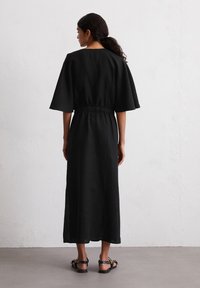 Abito di lino nero con maniche corte e larghe, vita cinchata e orlo fino alla caviglia. Il modello indossa sandali neri. Sfondo semplice, texture minimale.