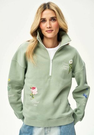 Kobieta o blond włosach nosi bluzę z suwakiem do ćwiartki w kolorze szałwii, ozdobioną kwiatowymi nadrukami i napisami takimi jak "Blush rose" i "Grain between paths."