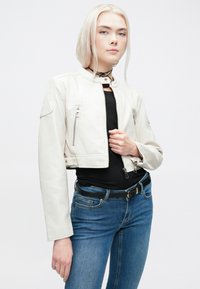 Lühendatud valge nahkjakk, millel on seisva kraega, esi- jahtkottide ning siledast tekstuurist, kantud musta särgi ja siniste teksade peal.