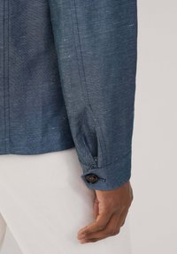 Ciemnoniebieska lniana koszula z zapięciem na guzik przy mankietach, charakteryzująca się teksturowanym splotem i dopasowanym krojem. Widoczne szwy podkreślają detale rękawa.