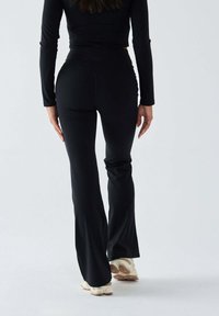 Czarne rozkloszowane spodnie treningowe wykonane z elastycznej tkaniny, zestawione z jasnymi butami sportowymi. Charakteryzują się wysokim pasem i gładką fakturą.