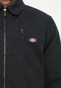 Chaqueta negra con cremallera, hecha de un tejido resistente, que presenta un cuello plegable, un bolsillo en el pecho con cremallera y una etiqueta con el logo de Dickies.
