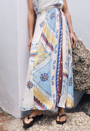 Femme portant une longue jupe avec des motifs de boussole et de vagues, debout pieds nus sur un sol rocheux près d'un mur blanc.