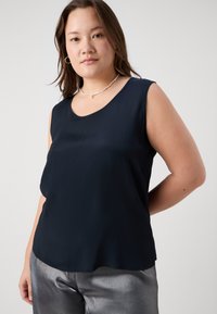 Blusa senza maniche blu navy in tessuto liscio con scollatura rotonda, abbinata a pantaloni metallici argento; delicato lucido e vestibilità rilassata.