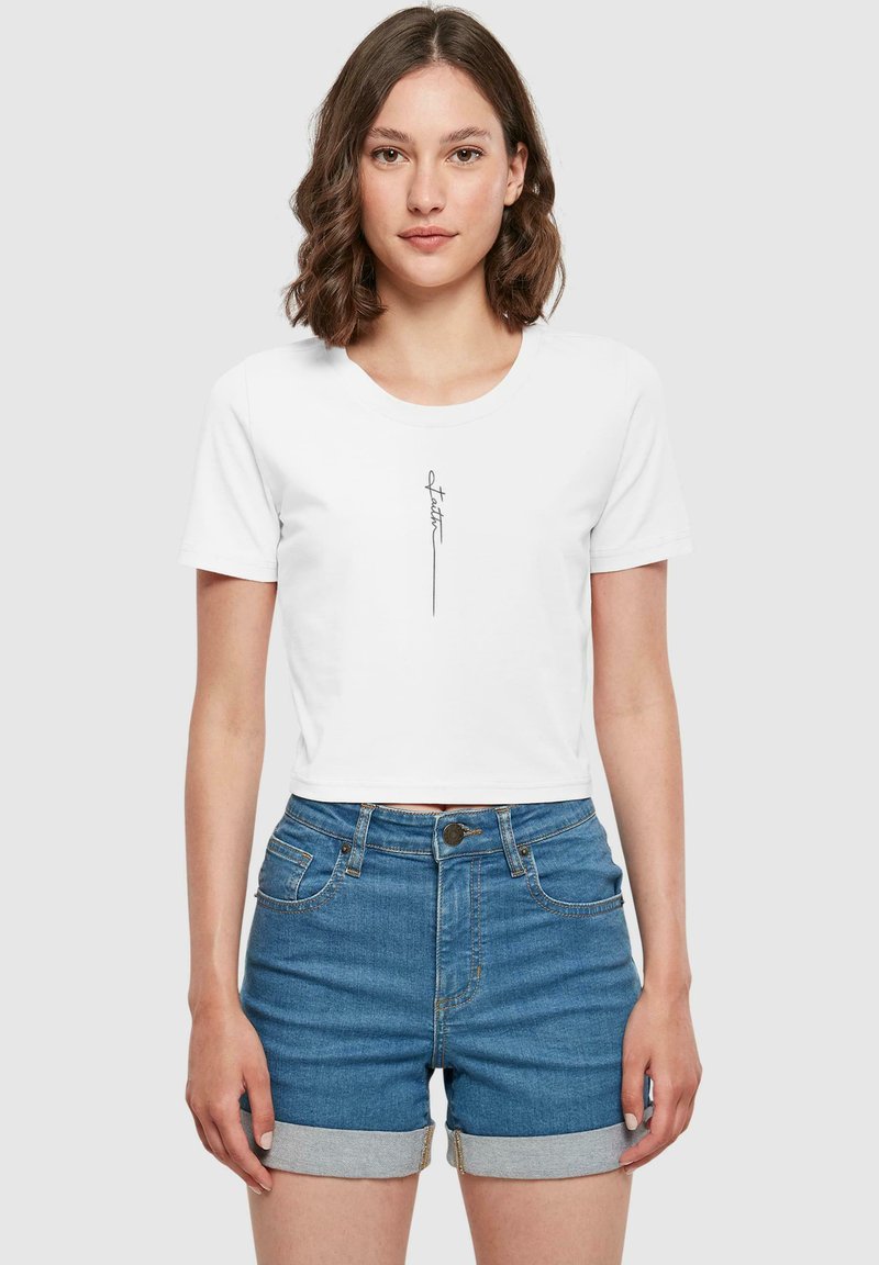 Camiseta blanca cropped de algodón, con un dibujo lineal minimalista en negro en la parte delantera. Combinada con pantalones cortos de mezclilla azul, con los dobladillos enrollados.