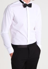 Mężczyzna w białej koszuli formalnej z czarnym muszką i czarnymi spodniami, z rękami w kieszeniach, przycięty na szyi i w talii.