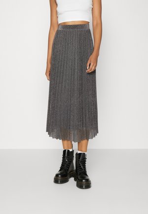 Femme portant une jupe midi plissée argentée, un débardeur court blanc, des chaussettes blanches et des bottines de combat noires à lacets sur fond blanc.