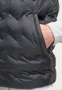 Chaqueta de plumas acolchada negra con un bolsillo lateral y una textura suave y brillante. El patrón en forma de flecha en la tela añade interés visual.