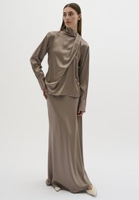 Satin brun langærmet top hænger elegant over en matchende maxi nederdel, der har høj hals og overdimensionerede manchetter, parret med spidse hvide sko.