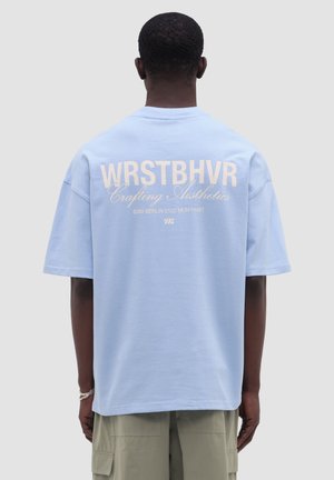 Mann trägt hellblaues WRSTBHVR T-Shirt und beige Cargo-Shorts, steht mit dem Rücken zur Kamera vor einfarbigem grauem Hintergrund.