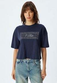 Camiseta recortada de color marino con mangas cortas, que presenta un diseño de estrellas y el texto "Girl Boss" en el centro. Textura de tela suave.