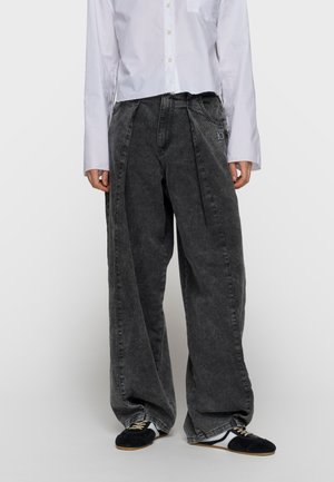 Person iført oversize vaskede sorte jeans med sidelommer, hvid skjorte med knapper og sorte og hvide sneakers mod en ensfarvet baggrund.