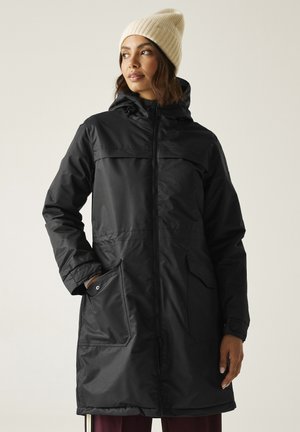 Mujer con gorro de punto beige que lleva un abrigo largo negro con capucha y cremallera, con grandes bolsillos delanteros, mirando hacia un lado.