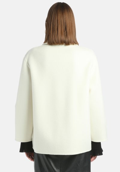 Valkoinen villasekoitekankaasta valmistettu ylipitkä neulepusero, jossa on pyöreä pääntie ja leveät hihat, mustilla ribbineulonnalla varustetut hihansuut. Teksturoitu pinta.