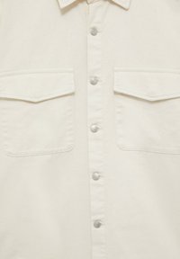 Camisa de botones color crema con cuello, dos bolsillos frontales y botones plateados. Hecha de una tela texturizada con un acabado duradero.