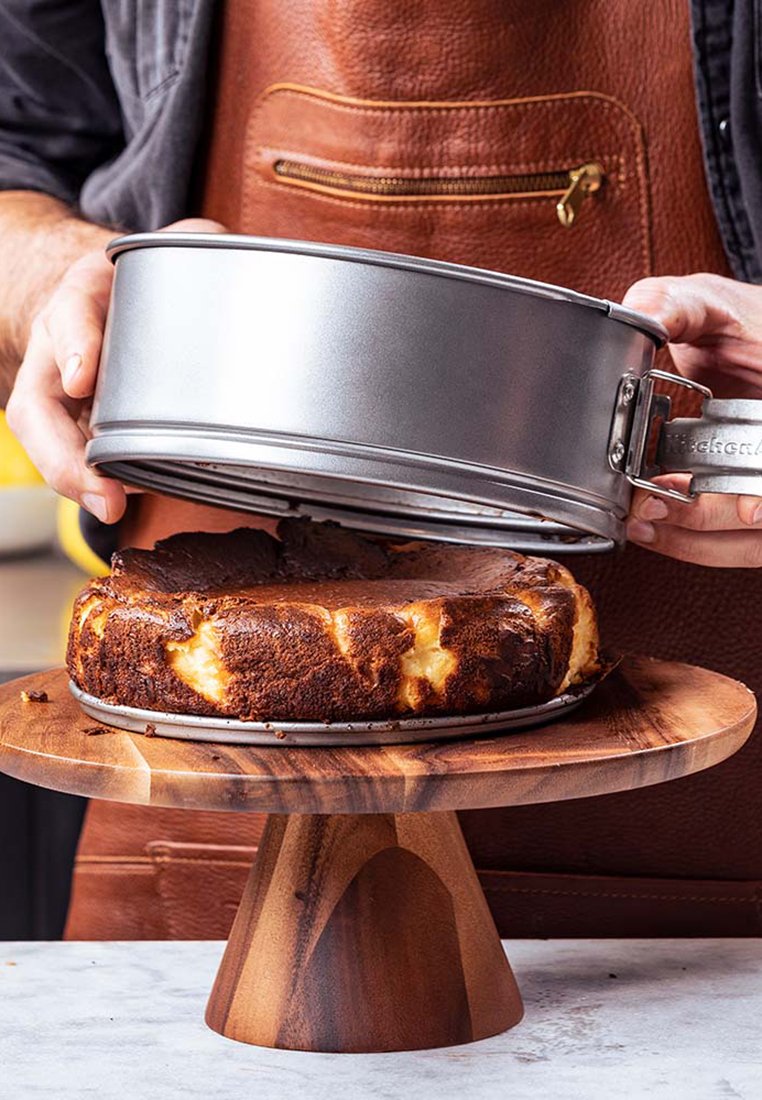 Ronde, goudbruin cake met een iets donkere rand staat op een houten voetstuk. Een zilveren springvorm wordt van de cake getild.