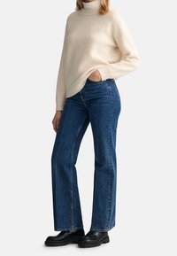 Suéter de cuello alto ribeteado color crema combinado con jeans acampanados de mezclilla azul de cintura alta y mocasines negros gruesos. Texturas lisas y silueta ajustada.