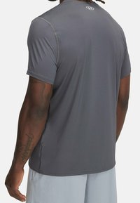 Hombre con camiseta deportiva de manga corta gris oscuro y pantalones cortos de malla gris claro, de pie con la espalda hacia la cámara.