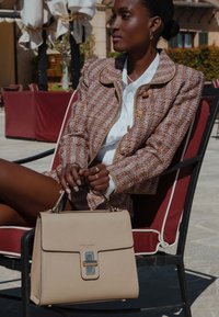 Beige Lederhandtasche mit strukturierter Form, ausgestattet mit einem silbernen Hardwareverschluss und einer texturierten Oberfläche, die neben einer Person in einem Tweedjackett liegt.