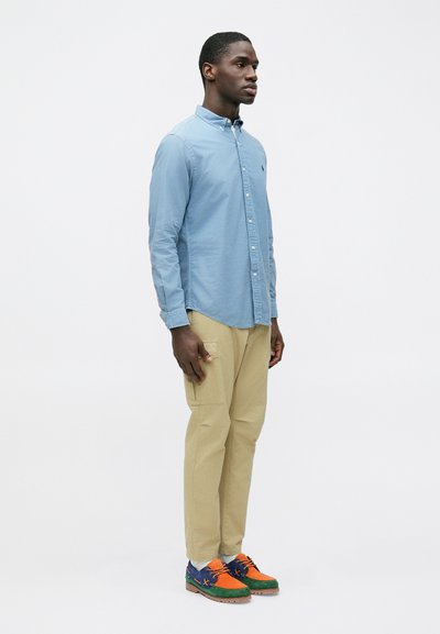 Camisa azul-clara com botões, calças cargo bege e sapatos multicoloridos (verde, laranja, azul) com superfície texturizada. Modelo em pé de lado.