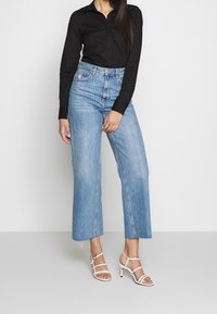 Chemise noire à boutons, jean clair taille haute à jambes larges, sandales à talons hauts blanches à lanières, tissu lisse, coupe droite, ourlets légèrement effilochés.