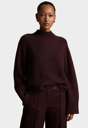 Femme portant un pull en tricot bordeaux foncé à manches longues avec des poignets et un col côtelés, associé à un pantalon taille haute assorti.