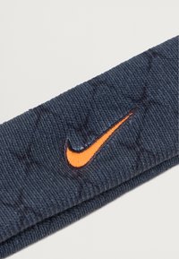 Námořnická látková čelenka s žebrovanou strukturou, s výrazným vyšitým logem Nike ve tvaru swoosh v oranžové barvě.