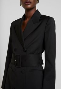 Blazer negro ajustado con un dise�ño de doble botonadura, cuello de muesca y un cinturón ancho. Cuenta con bolsillos laterales y botones en la parte frontal.