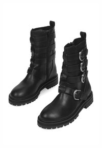 Botas de cuero negras hasta el tobillo con un diseño en capas, cierre de cremallera y tres hebillas plateadas. Suela gruesa con relieve para mayor durabilidad.