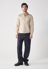 Camicetta beige con colletto e logo sul petto, abbinata a jeans blu scuro a vestibilità ampia e scarpe nere. Design e texture semplici.
