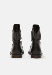 Botas de tobillo de cuero negro con una textura suave, puntas redondeadas, costuras decorativas y tacones bajos apilados. La vista muestra la parte posterior y los lados.