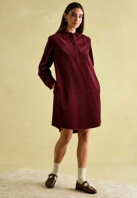 Bordeauxfarbiges Baumwollkleid mit hohem Kragen, Druckknopfblende, langen Ärmeln und seitlichen Taschen. Kombiniert mit braunen Schuhen und gepunkteten Socken.