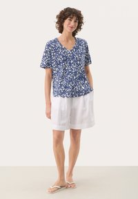 Blå och vit mönstrad kortärmad topp med V-ringning. Matcher med vita linneshorts. Modell som bär flip-flops. Enkel, avslappnad look.