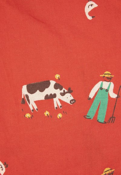 Contadino con tuta verde e cappello che tiene una forca vicino a una mucca bianca e nera circondata da fiori gialli su uno sfondo di tessuto rosso.