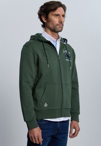 Sudadera con capucha verde con cremallera, capucha con cordón, bolsillos delanteros y detalles bordados. Presenta una textura suave y puños acanalados.
