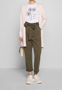 Cardigan rosa claro sobre una camiseta blanca con gráficos florales, pantalones de talle alto verde oliva con un lazo, zapatillas beige y un bolso pequeño.
