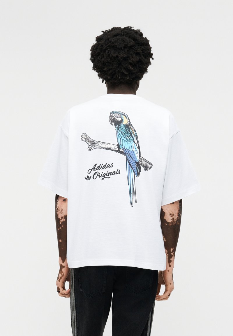 Person mit dunklem, lockigem Haar trägt ein weißes Adidas Originals T-Shirt mit einem blauen und gelben Papagei, der auf einem Ast auf dem Rücken sitzt.