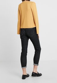 Gul, ribbad tröja med figurnära design och långa ärmar. Svarta smala jeans som är uppvikt vid fållarna. Svarta platta skor. Enhetlig bakgrund.