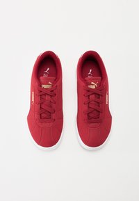 Røde sneakers med en glat tekstiloverdel, rund tå og hvit gummisåle. Snøring med gull logo-detaljer på siden.