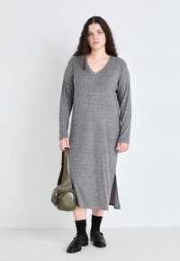 Rochie lungă, tricotată, de culoare gri, cu decolteu în V, mâneci lungi și slituri laterale. Asortată cu pantofi negri, ciorapi până la genunchi și o geantă de umăr de culoare verde măslinie.