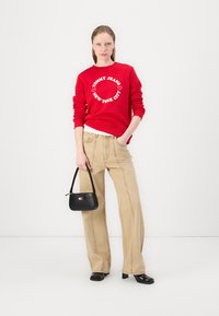 Rød hettegenser med hvit logo, beige vide bukser og en liten svart håndveske. Outfit'en har en uformell, avslappet passform.